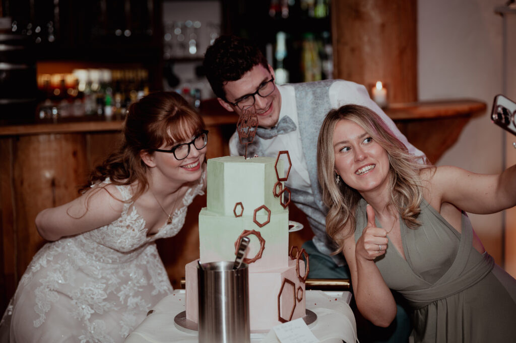 Brautpaar und Gästin posieren lachend vor der Hochzeitstorte im Hotel Backenköhler in Ganderkesee, fotografiert von Hochzeitsfotograf Björn Stuhr.